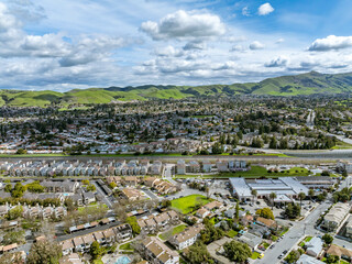 Drone images showcasing residential neighborhoods in Fremont, California, with lush green hills in the background and a mix of townhomes, single-family homes, and commercial buildings. Ideal for theme