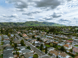 Drone images showcasing residential neighborhoods in Fremont, California, with lush green hills in the background and a mix of townhomes, single-family homes, and commercial buildings. Ideal for theme