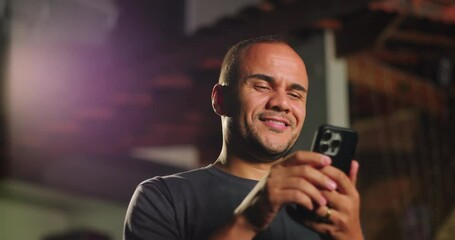 Latin Hispanic man of African descent smiling at smartphone while enjoying moment outdoors, reacting with genuine amusement to something funny seen on screen