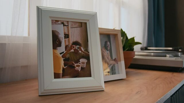 Close-up of framed family photos on cabinet in living room of family home, next to vinyl record player, depicting African American father and son having fun with doughnuts while drinking tea