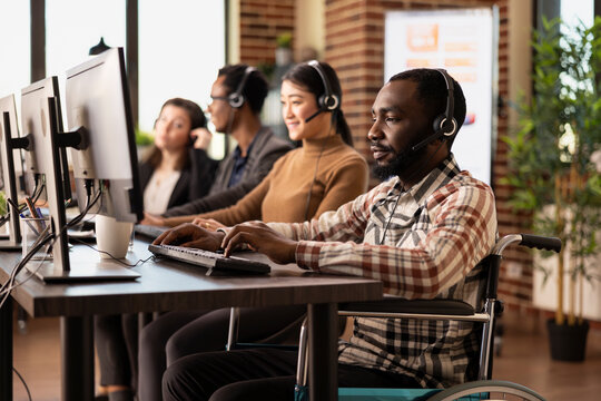 Black male wheelchair user with headset, engaged in phone call with client and reviewing details on desktop pc. Businessman with physical injury, sitting among his coworkers and using his computer.