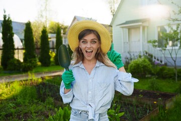 Portrait of a smiling gardener woman standing next to her green garden holding a garden spatula,...