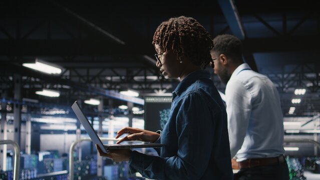 Data center computer scientist doing maintenance tasks on laptop, ensuring optimal performance. African american technician reviewing diagnostics and applying patches to server hub gear,