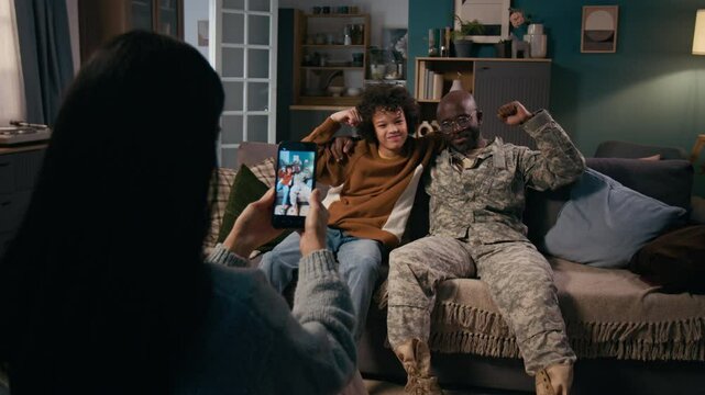 Medium full rear shot of young wife taking photographs of diverse African American military officer husband and polyethnic son relaxing together on couch, hugging, posing with flexed biceps