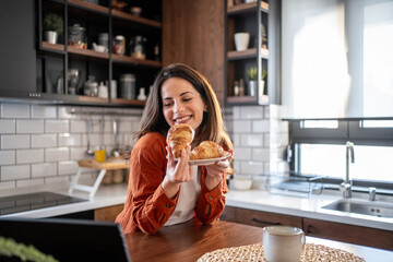Happy woman showing croissants in video call from kitchen table