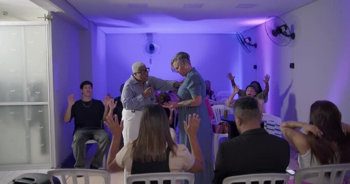 Female preacher praying over another woman during evangelical church service as seated congregation lifts hands in unified act of worship and spiritual support