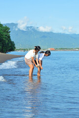 Two girls bend toward the shallow beach water, closely looking at something interesting beneath the...
