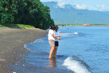 Standing at the water's edge, two girls examine something they've picked up from the beach, framed...