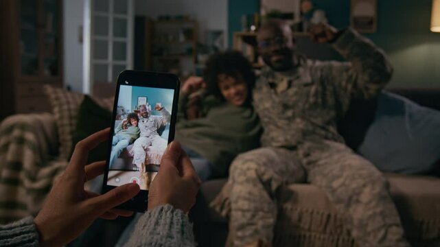 Medium shot of hands of anonymous woman taking photos of African American marine husband and biracial son hugging together, posing, flexing biceps on couch, boy happy at father returning home