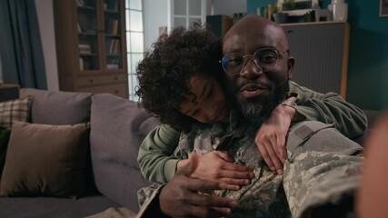 Medium close-up POV of cheerful African American army soldier in khaki military uniform sitting on couch at home, chatting on video call, smiling, looking at camera, hugging teenage son