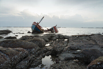 Shipwrecked fishing boat stranded on rocky coast under dramatic cloudy sky, symbolizing danger at...