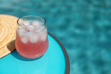 Refreshing pink beverage served in a glass filled with ice, on a turquoise table next to a straw hat, with a blurred blue swimming pool in the background with a space for text. summer vacation scene