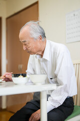 In a modest Japanese apartment, a man in his late 70s wearing a white shirt sits at a small table, eating a bowl of natto rice for breakfast as soft morning light filters gently through the window.
