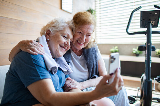 Senior lesbian couple laughing while looking at smartphone after home workout - Powered by Adobe