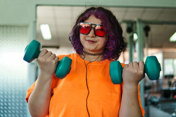 Beautiful young overweight Latina posing and exercising with weights and listening to music, wearing retro glasses and a Discman in a retro-style gym, working hard, very happy. Health and improvement.