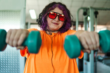 Beautiful young overweight Latina posing and exercising with weights and listening to music, wearing retro glasses and a Discman in a retro-style gym, working hard, very happy. Health and improvement.