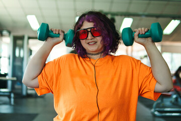 Beautiful young overweight Latina posing and exercising with weights and listening to music, wearing retro glasses and a Discman in a retro-style gym, working hard, very happy. Health and improvement.