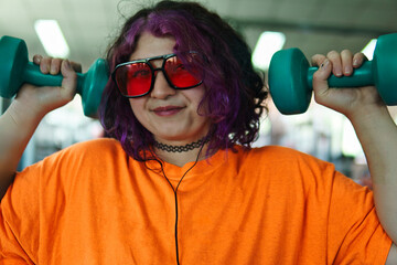 Beautiful young overweight Latina posing and exercising with weights and listening to music, wearing retro glasses and a Discman in a retro-style gym, working hard, very happy. Health and improvement.