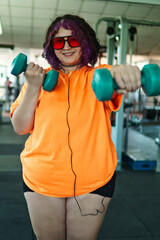 Beautiful young overweight Latina posing and exercising with weights and listening to music, wearing retro glasses and a Discman in a retro-style gym, working hard, very happy. Health and improvement.