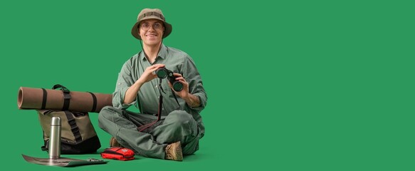 Male traveler with binoculars and travel accessories sitting on green background with space for text