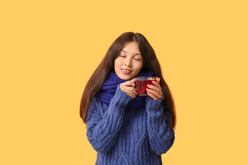 Beautiful young happy Asian woman in winter clothes with cup of hot mulled wine on yellow background