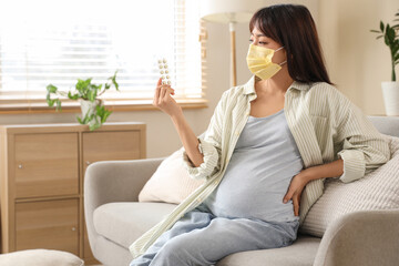 Pregnant Asian woman in yellow medical mask with pills sitting on sofa at home