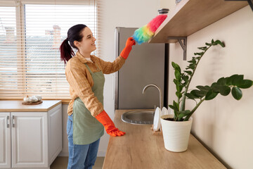 Young woman with pp-duster cleaning shelf in kitchen