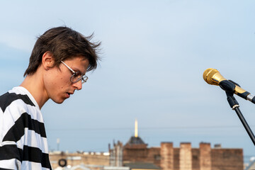 A young caucasian male in striped attire stands next to a microphone on a rooftop. the blurred urban backdrop and clear sky provide a vibrant setting, capturing creativity and urban life.