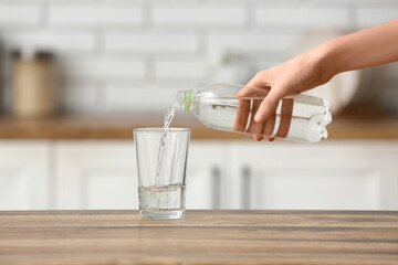 Woman pouring water from bottle into glass on wooden table in kitchen, closeup