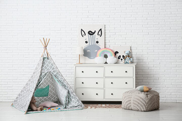 Interior of children's room with play tent, many toys and lamp on commode near white brick wall
