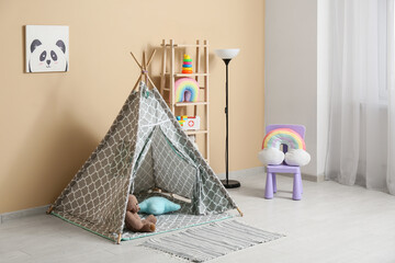Interior of children's room with play tent, lamp, chair and many toys in shelving unit near beige wall