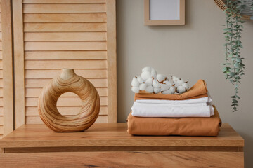 Stack of clean bed sheets with cotton flowers sprig and vase on commode near beige wall