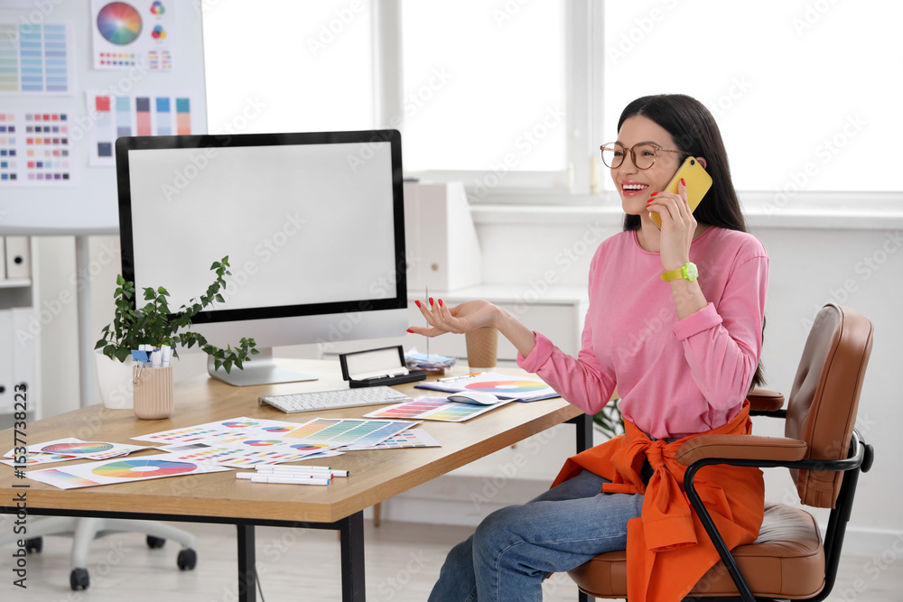 Fototapete female designer with blank computer talking by mobile phone at table in office