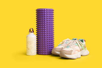 Foam roller with bottle of water and stylish sneakers on yellow background