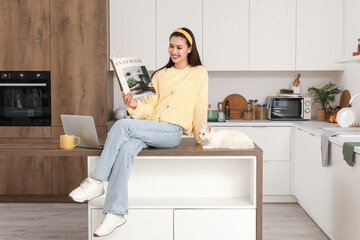 Beautiful young happy woman with cute white cat reading magazine on table in kitchen