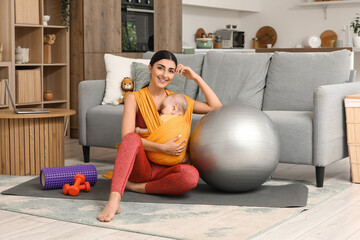Sporty mother with her baby in sling and fitball sitting at home