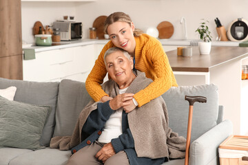 Young caregiver hugging senior woman with plaid on sofa in kitchen