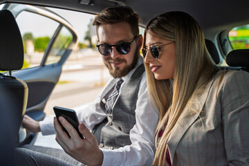 Elegant young man and woman in suits looking at smartphone in the back of the car 