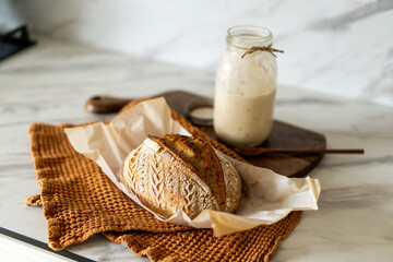 Crispy sourdough bread sits beside a glass jar filled with active starter, accompanied by a rustic...