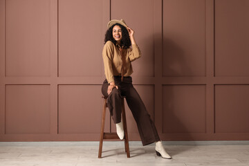 Stylish young African-American woman in hat sitting on stool near brown wall