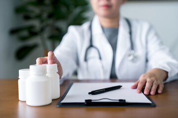 Close-up of doctor with stethoscope reaching for medication bottle beside clipboard, highlighting patient prescription management and written treatment planning in healthcare settings.