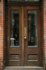 A detailed close-up of a vintage wooden double door with glass inserts, set in a red brick wall. The craftsmanship features carved trim and a brass handle with classic charm.