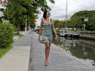 Beautiful Asian woman walks along a paved canal path, wearing a green floral dress with ruffled...