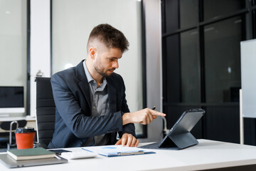 A smart Caucasian businessman sits confidently at desk in modern indoor office, using laptop for online consulting, showcasing professionalism, success, digital expertise in corporate environment.