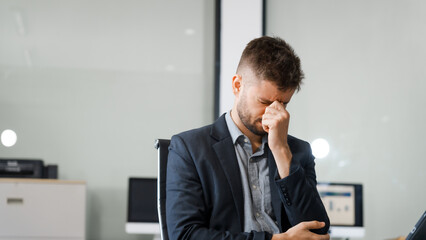 A stressed Caucasian businessman in a suit sit indoors at an office desk, suffering from office...
