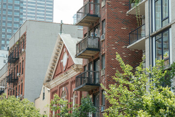 A red-brick church roof with classical trim stands beside old and new apartment buildings. Balconies, fire escapes, and high-rises contrast in this layered urban cityscape.