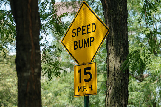 A bright yellow speed bump sign with a 15 MPH limit stands among green trees. The sign is clearly visible, warning drivers in a shaded, wooded neighborhood.
