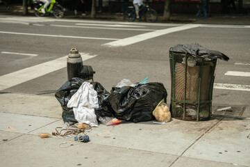 Black garbage bags and loose trash lie next to an overflowing street bin on a concrete sidewalk. A...