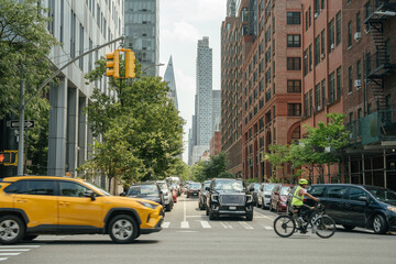 A cyclist in a reflective vest rides through a busy New York street surrounded by cars and tall buildings. A yellow SUV crosses the intersection while modern skyscrapers rise in the background.