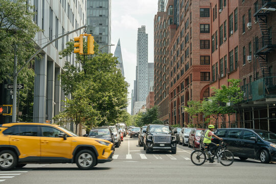 A cyclist in a reflective vest rides through a busy New York street surrounded by cars and tall buildings. A yellow SUV crosses the intersection while modern skyscrapers rise in the background.
